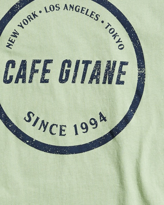 The Cafe Gitane classic t-shirt in green is printed with the logo in a stamped structure. The print ins circular in the middle of the front chest and in navy blue. The logo features each city Cafe Gitane has a location in: New York, Los Angeles, and Tokyo, as well as when the first location opened: 1994.  Edit alt text