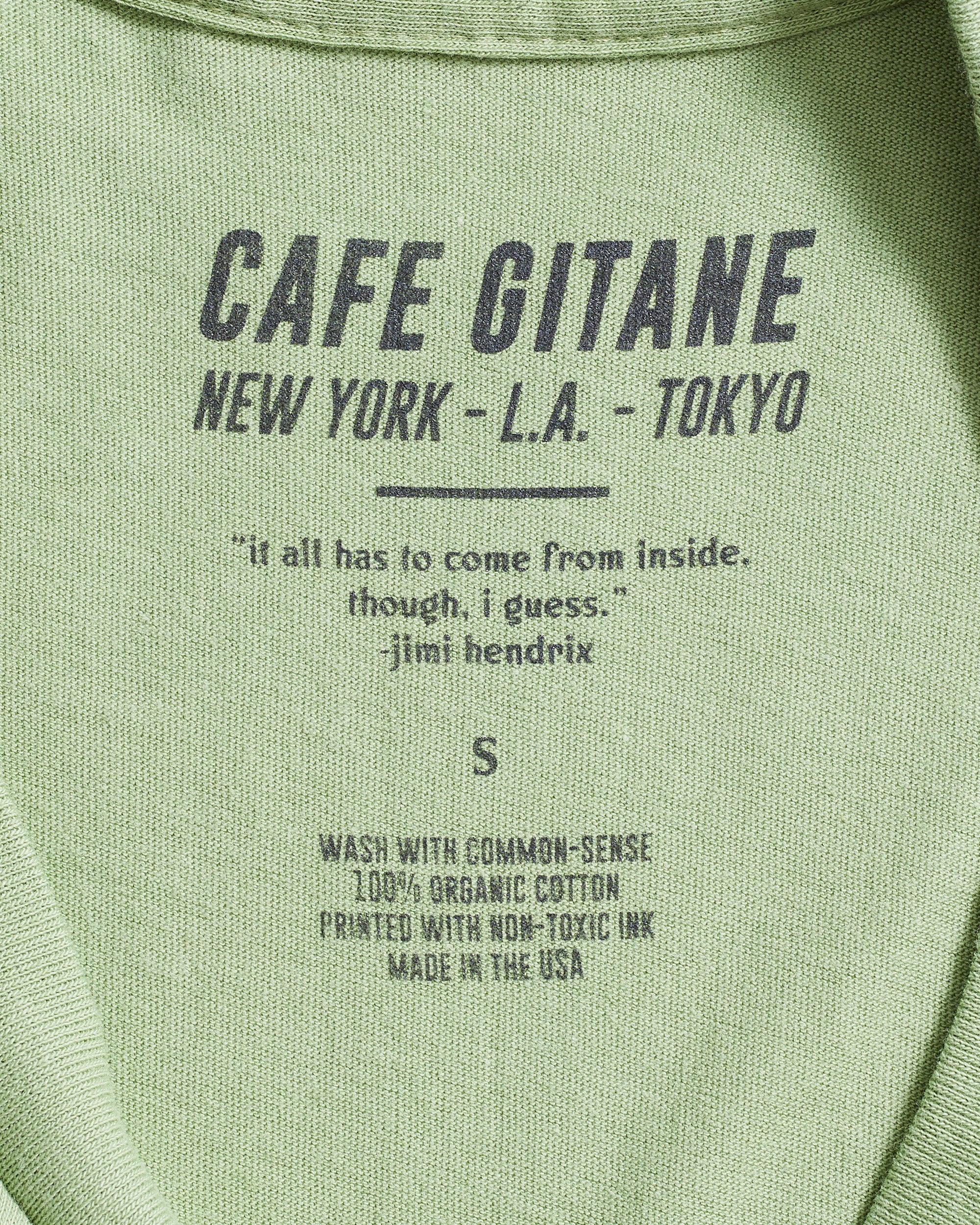 The Cafe Gitane 100% organic cotton t-shirt in green has a printed inside tag. It show the care instruction, that it is made in the USA and includes a Jimi Hendrix Quote.  It also has the Cafe Gitane logo and stated the cities it has locations in: New York, Los Angeles, and Tokyo. 