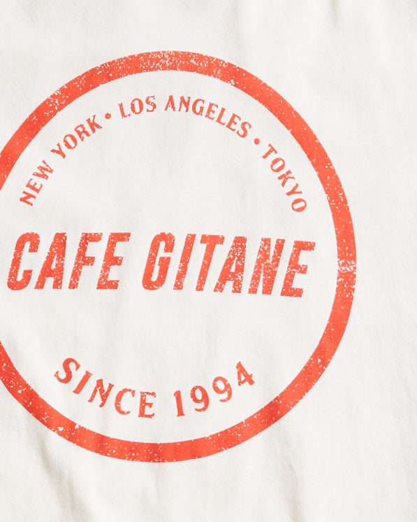 The Cafe Gitane Stamped Logo Tee is 100% organic cotton and made in the USA. The classic t-shirt in natural white and printed with a red logo in a stamped structure. The print ins circular in the middle of the front chest. The logo features each city Cafe Gitane has a location in: New York, Los Angeles, and Tokyo, as well as when the first location opened: 1994. 