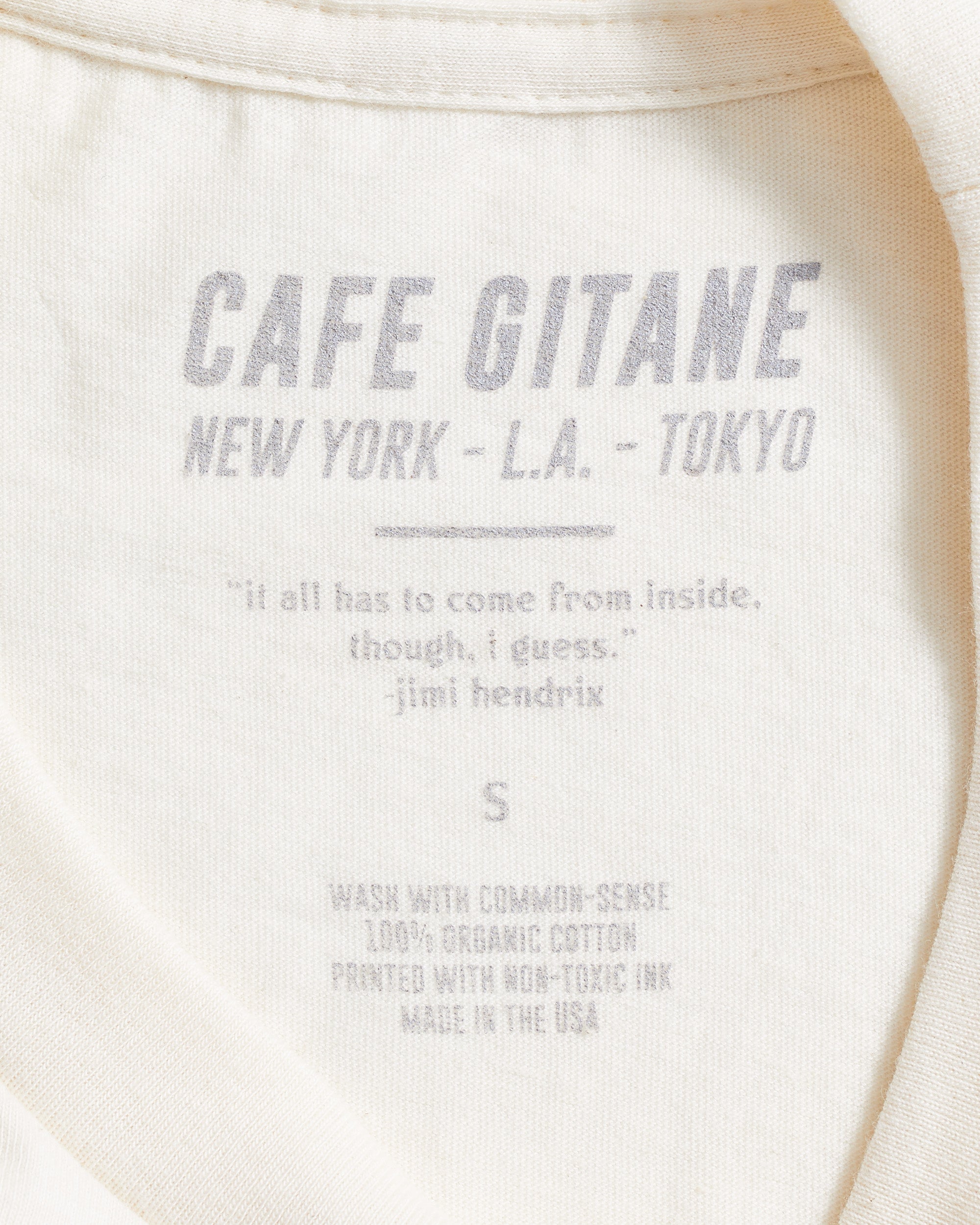 The Cafe Gitane 100% organic cotton t-shirt in green has a printed inside tag. It show the care instruction, that it is made in the USA and includes a Jimi Hendrix Quote.  It also has the Cafe Gitane logo and stated the cities it has locations in: New York, Los Angeles, and Tokyo. 