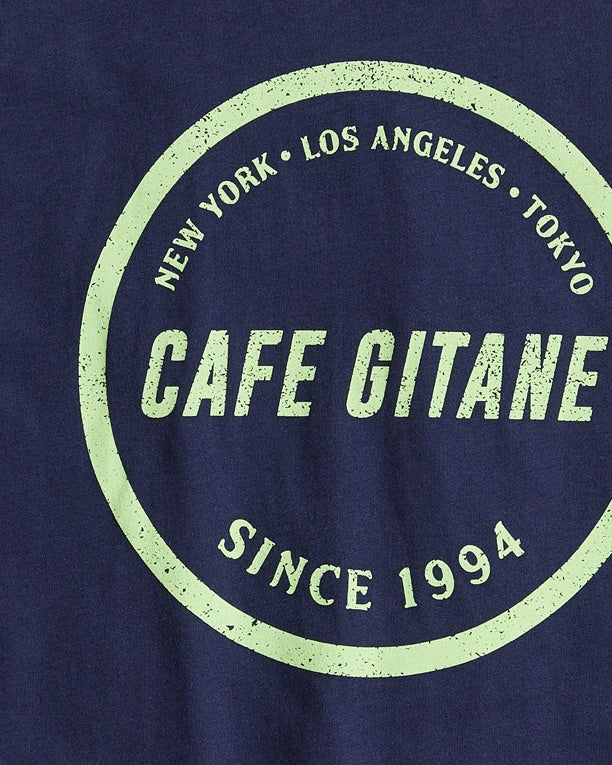 The Cafe Gitane Stamped Logo Tee is 100% organic cotton and made in the USA. The classic t-shirt in navy blue and printed with a green logo in a stamped structure, with non-toxic ink. The print ins circular in the middle of the front chest. The logo features each city Cafe Gitane has a location in: New York, Los Angeles, and Tokyo, as well as when the first location opened: 1994. 