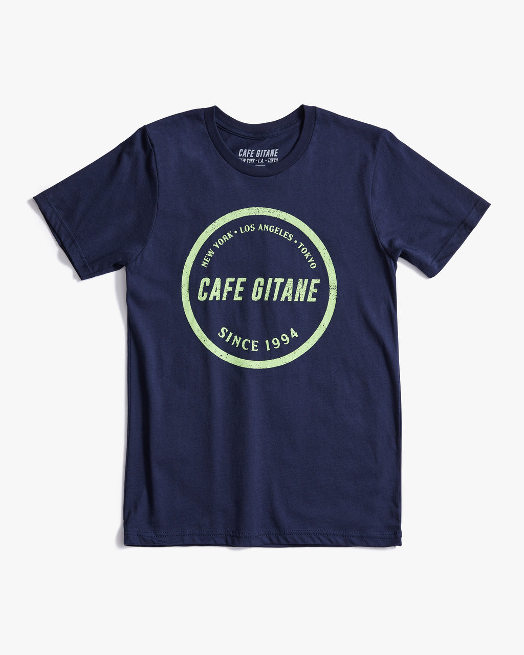 The Cafe Gitane Stamped Logo Tee is 100% organic cotton and made in the USA. The classic t-shirt in navy blue and printed with a green logo in a stamped structure, with non-toxic ink. The print ins circular in the middle of the front chest. The logo features each city Cafe Gitane has a location in: New York, Los Angeles, and Tokyo, as well as when the first location opened: 1994. 