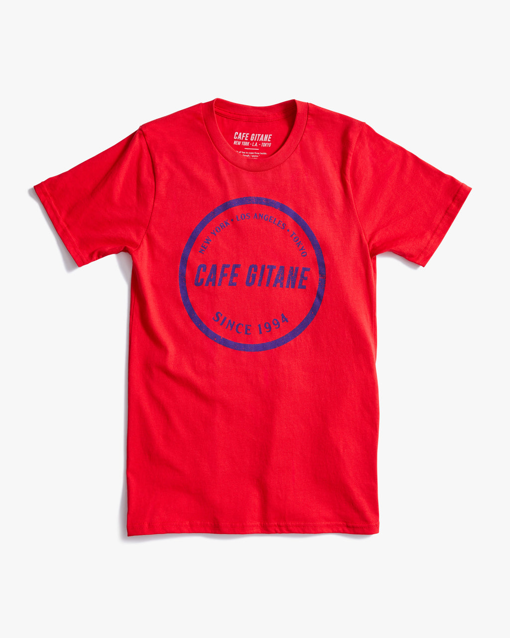 The Cafe Gitane Stamped Logo Tee is 100% organic cotton and made in the USA. The classic t-shirt in red and printed with a royal blue logo in a stamped structure, with non-toxic ink. The print ins circular in the middle of the front chest. The logo features each city Cafe Gitane has a location in: New York, Los Angeles, and Tokyo, as well as when the first location opened: 1994. 