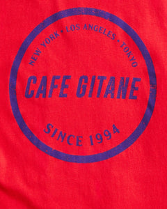 The Cafe Gitane Stamped Logo Tee is 100% organic cotton and made in the USA. The classic t-shirt in red and printed with a royal blue logo in a stamped structure, with non-toxic ink. The print ins circular in the middle of the front chest. The logo features each city Cafe Gitane has a location in: New York, Los Angeles, and Tokyo, as well as when the first location opened: 1994.  Edit alt text