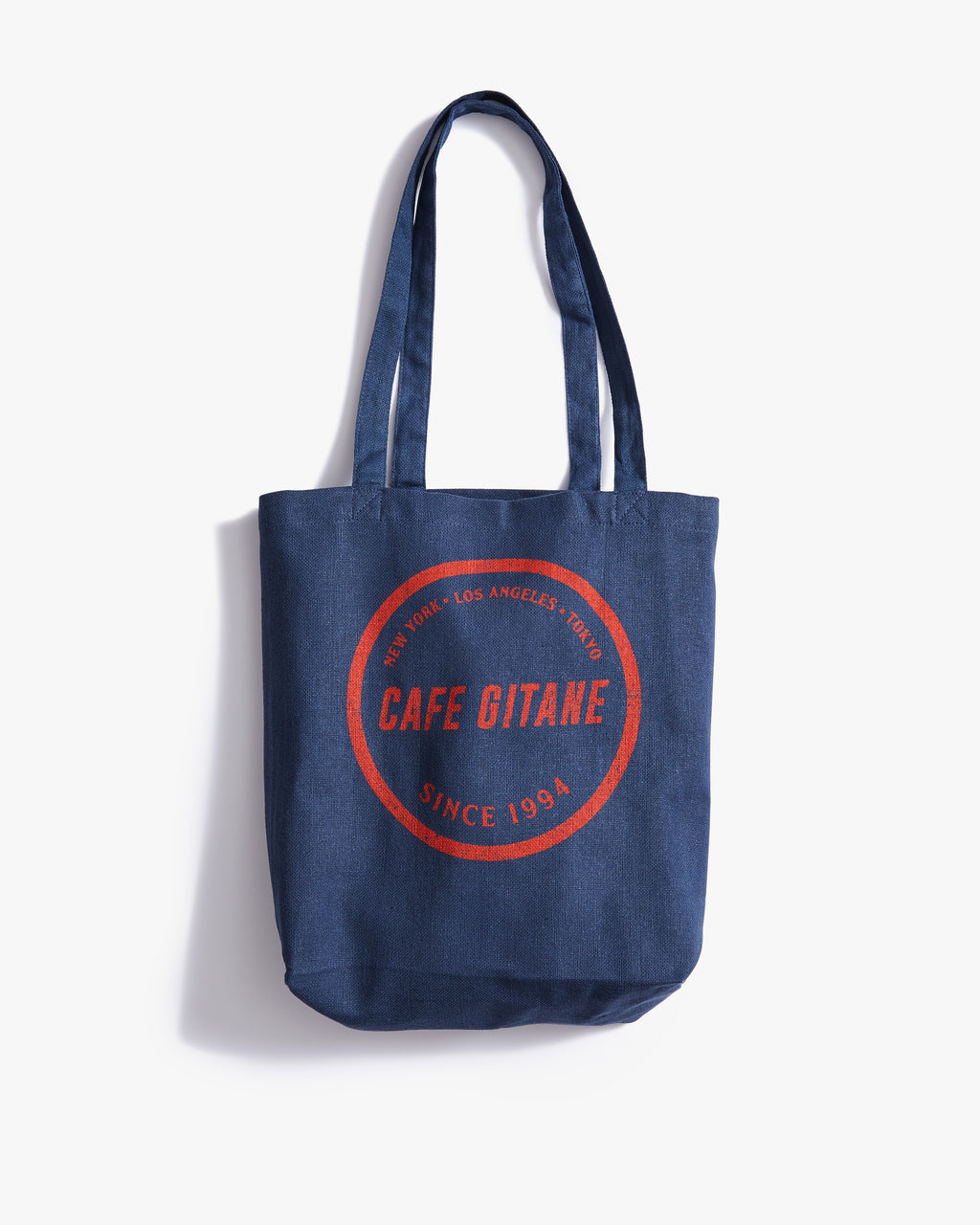 The Cafe Gitane Stampe Logo Tote Bag is 100% recycled cotton and made in the USA. The classic market bag is navy blue and printed with a red logo in a stamped structure, with non-toxic ink. The logo features each city Cafe Gitane has a location in: New York, Los Angeles, and Tokyo, as well as when the first location opened: 1994. 