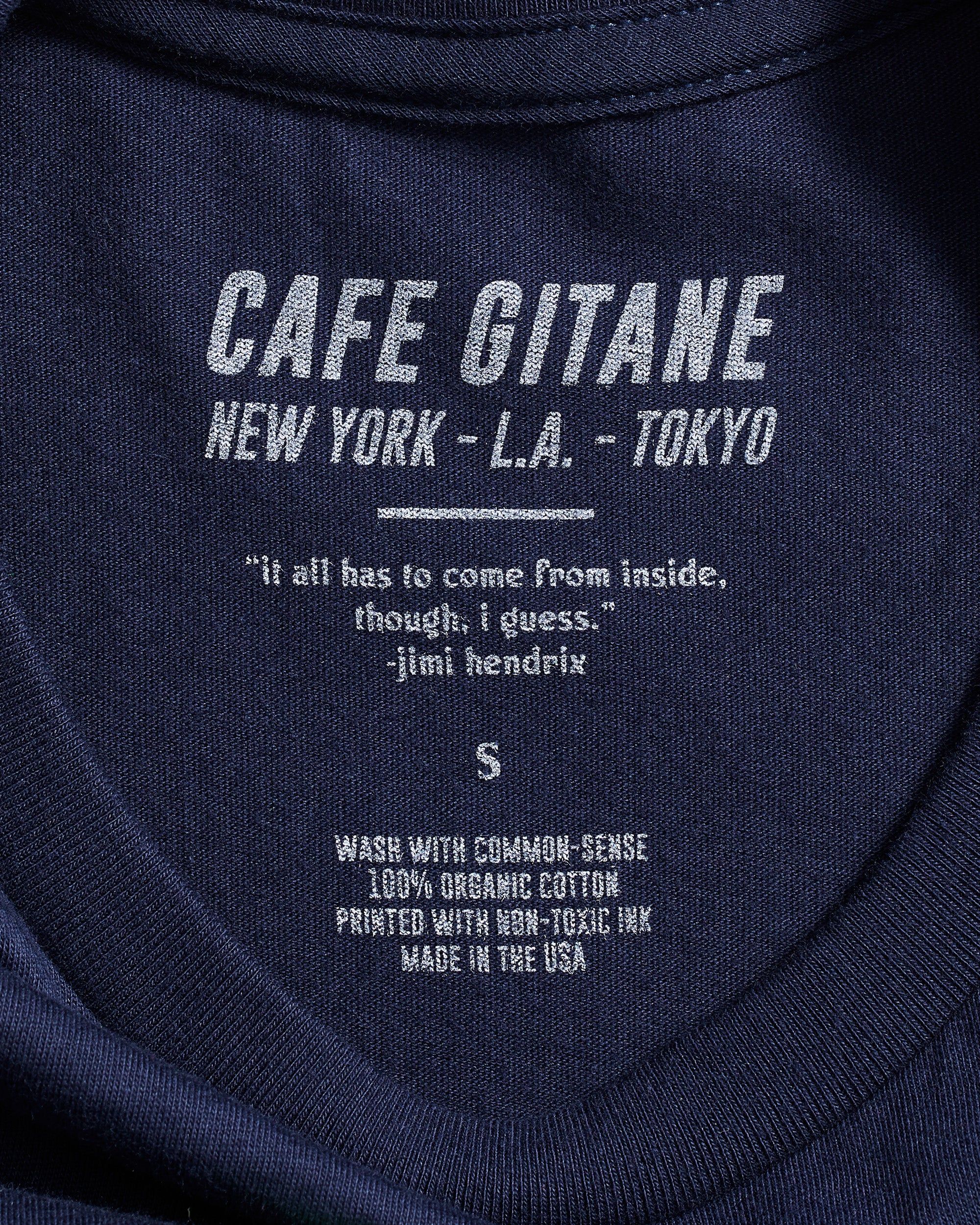 The Cafe Gitane 100% organic cotton t-shirt in navy blue has a printed inside tag. It show the care instruction, that it is made in the USA and includes a Jimi Hendrix Quote.  It also has the Cafe Gitane logo and stated the cities it has locations in: New York, Los Angeles, and Tokyo. 